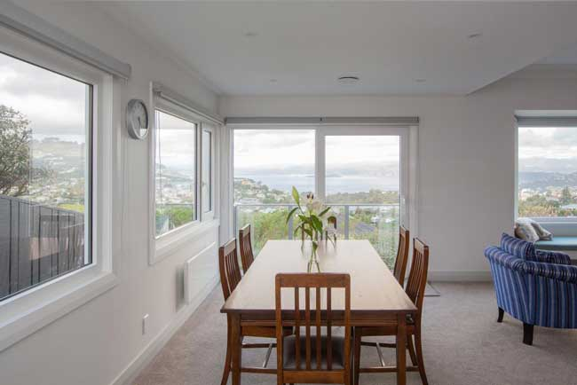 Dining room with a wooden table and chairs, large windows with clean uPVC window frames offering a scenic view of water and hills, minimalistic decor, and a blue-striped armchair in the corner.