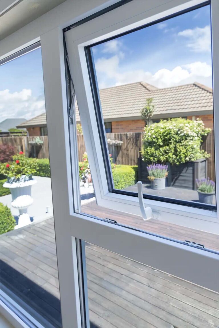Tall white uPVC awning window shown open, looking toward the garden and neighbouring brick house.